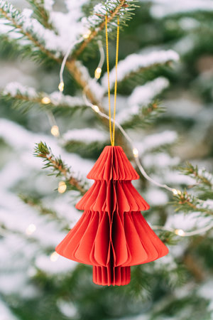 Red paper christmas tree ornament hanging on snow-covered pine with lights. Festive composition, mockup for a New Years cardの写真素材