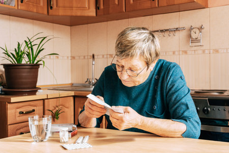 An elderly woman reads pill instructions in kitchen setting. Health maintenance, healthy aging, vitamin support, home treatmentの写真素材