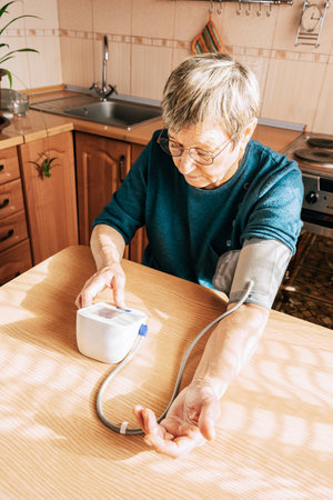 An elderly woman measures her blood pressure in the kitchen. Health maintenance, healthy aging,...