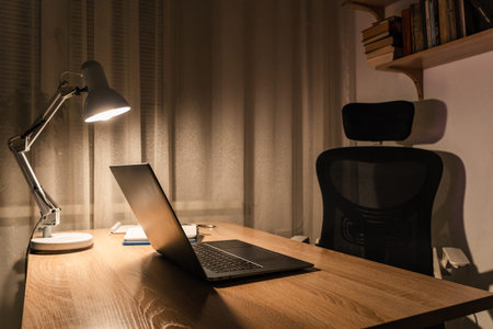 Workspace with laptop, illuminating desk lamp, empty chair, and books on shelf, symbolizing...
