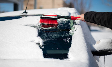 A young man wearing black warm suit for cleans his car after a snowfall on a sunny, frosty day....