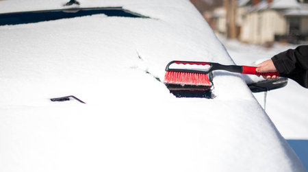 A young man wearing black warm suit for cleans his car after a snowfall on a sunny, frosty day. Cleaning and clearing the car from snow on a winter day and a severe snowstorm in winter.の写真素材