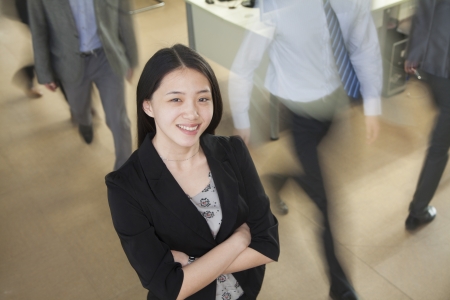 Young businesswoman smiling in the office, businessmen walking all around herの写真素材