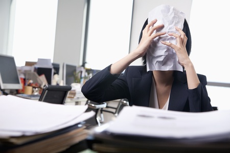 Young businesswoman sitting at desk covering her face with a paperの写真素材