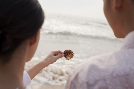 Young couple standing and looking at seashell on the beach, over the shoulder viewの写真素材