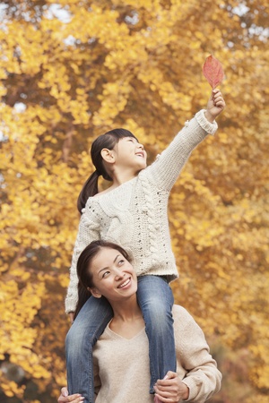 Daughter Riding Piggy-Back on Mother's Shoulders Enjoying Autumn Leavesの写真素材
