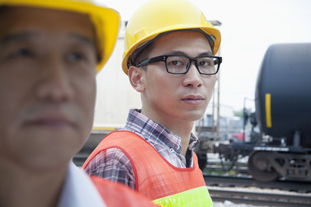 Serious engineer in protective workwear looking at camera outside in front of railroad tracks 