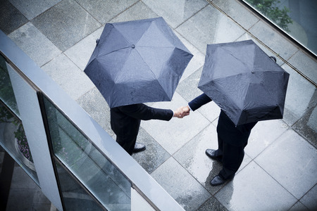 High angle view of two businessmen holding umbrellas and shaking hands in the rainの写真素材