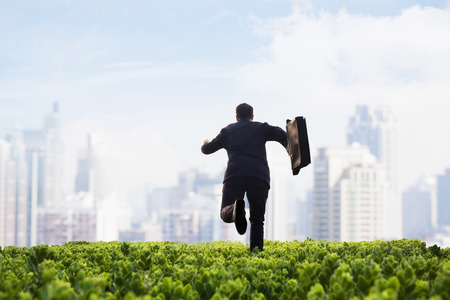 Businessman running towards the city with a briefcase in a green field with plantsの写真素材