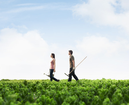 Two young people carrying gardening equipment walking across a green field with plantsの写真素材