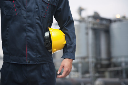 Midsection of young worker holding a yellow hardhat  outdoors with factory in the background