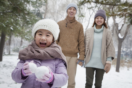 Young girl carrying snow balls in front of parents in park in winterの写真素材