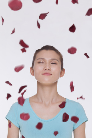 Beautiful tranquil young woman looking up with eyes closed with rose petals flying around herの写真素材