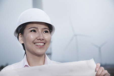 Young smiling female engineer holding a blueprint with wind turbines in the backgroundの写真素材