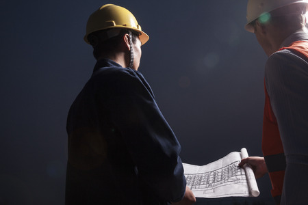 Two young male engineers in hardhats looking down at a blueprint at dusk, rear viewの写真素材