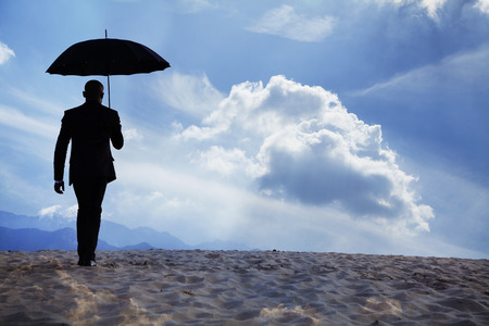 Businessman holding an umbrella and walking away in the middle of the desert with dreamlike cloudsの写真素材