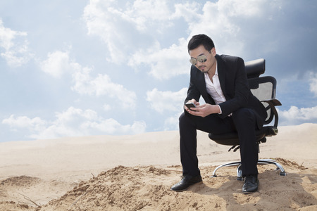 Young businessman in sunglasses sitting in an office chair in the middle of the desert, checking his phoneの写真素材