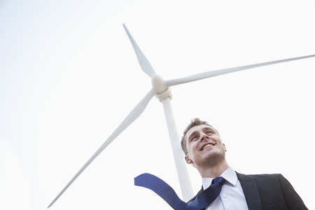 Young smiling businessman standing beside a wind turbine, tie is blowing in the windの写真素材