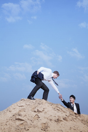 Young businessman helping another businessman get to the top of the hill in the desertの写真素材