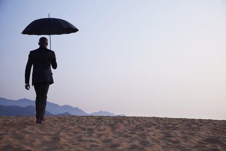 Businessman holding an umbrella and walking away in the middle of the desertの写真素材