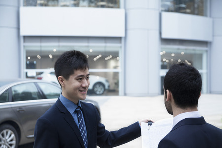 Car salesman holding car keys and paperwork and selling a car to a young businessmanの写真素材