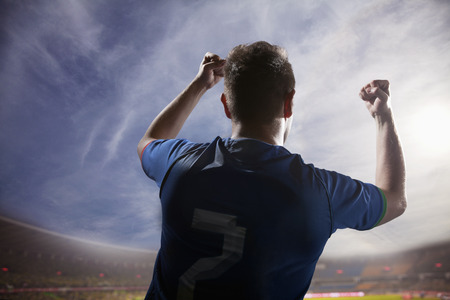 Soccer player with arms raised cheering, stadium with sky and cloudsの写真素材