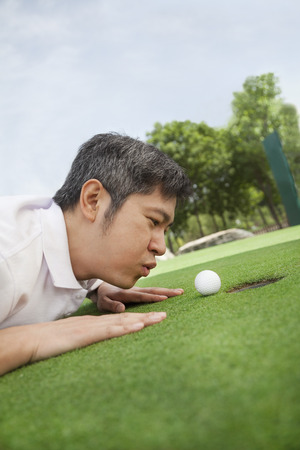 Mid adult man lying down in a golf course trying to blow the ball into the holeの写真素材