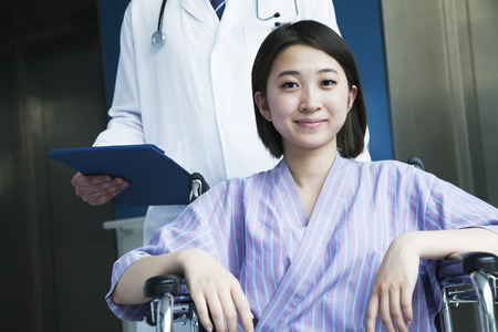 Young smiling female patient sitting in a wheelchair, doctor standing behind her, looking at cameraの写真素材