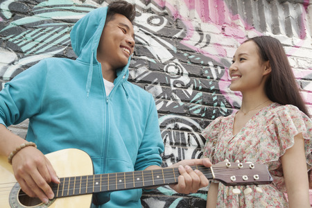 Young smiling street musician leaning on a wall with graffiti drawings, playing his guitar, and flirting with a young woman in a dressの写真素材
