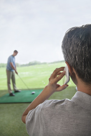 Rear view of man on the phone while another man plays golf in the backgroundの写真素材