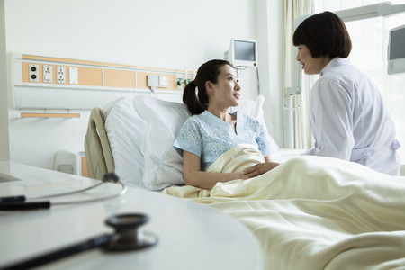Female doctor sitting on hospital bed and discussing with young female patientの写真素材