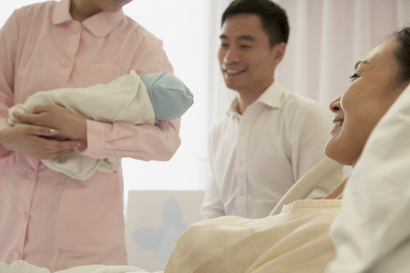 Nurse holding newborn baby in the hospital with mother lying on the bed and the father beside herの写真素材