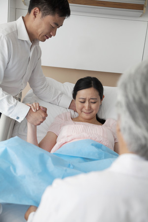 Women in labor holding her husbands hand with doctor in the foreground in the hospitalの写真素材