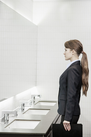 Young businesswoman with long hair looking into the mirror and preparing herself in the bathroomの写真素材