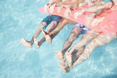 Low section of four friends in a pool holding onto an inflatable raft with feet sticking out of the...