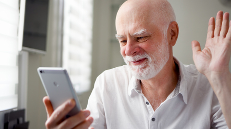 Senior man sitting at home with smartphone. Using mobile talking via messenger app Skype. Smiling waving hand in greeting. Remote freelance work on retirement, active modern lifestyle of older peopleの写真素材