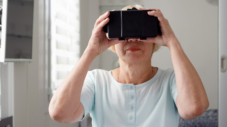 Good-looking blond senior woman in white using VR 360 glasses at home. Concept of active modern elderly people, learning to use contemporary technologies.の写真素材