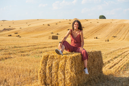 Pretty and smiling young girl with curly hair. Sitting on a straw bale in a wheat field.の写真素材