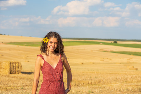 Pretty young girl with curly hair. Smiling and with a sunflower in her hair. In a rural setting. With a wheat field in the background.の写真素材