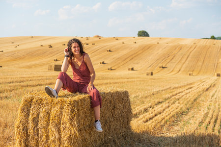 Pretty and smiling young girl with curly hair. Sitting on a straw bale in a wheat field.の写真素材