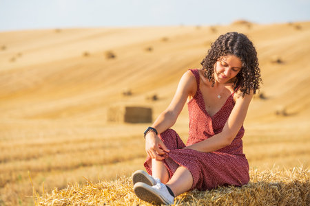 Pretty and smiling young girl with curly hair. Sitting on a straw bale in a wheat field.の写真素材