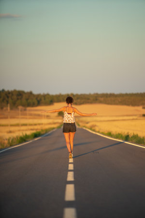 Young girl, walking in the middle of a lonely straight road, late in the day. With the hands stretched out in the shape of a cross, to maintain balance and not fall from the dividing line.の写真素材