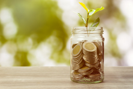 Coins in glass jar on wooden table with green bokeh background, saving money concept