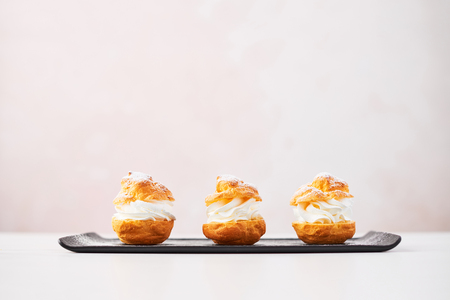 Delicious cream puff cakes with cream and powdered sugar on a black plate on white marble table over pink background. Selective focus, copy space.の写真素材