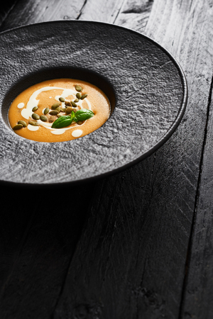 Pumpkin and carrot soup with pumpkin seeds, cream and basil in black modern plate on dark wooden table with copy space. Dark modern style.の写真素材