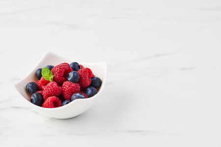 Fresh blueberries and raspberries with mint leaf in white bowl on white marble table with copy space.の写真素材