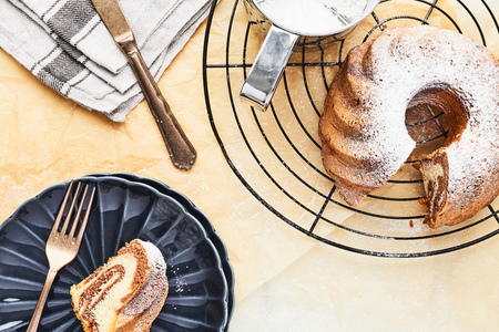 Marble bundt cake on french cooling rack with powdered sugar and one slice of cake on dark blue side plate over baking paper. Holiday baking background. Top view with copy space for text.の写真素材