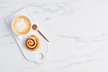 Freshly baked cinnamon bun with spices and cocoa filling and coffee or cappuccino with latte art on white serving plate over white marble background. Top view. Copy space for text. Swedish breakfast.の写真素材