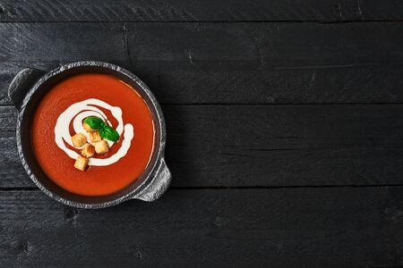 Tomato soup with cream, croutons and basil in a black bowl on wooden background. Top view. Copy...