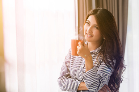 Asian woman in her living room drinking holding a coffee tea mug with sunrise streaming in through window and creating flare into the lens.の写真素材
